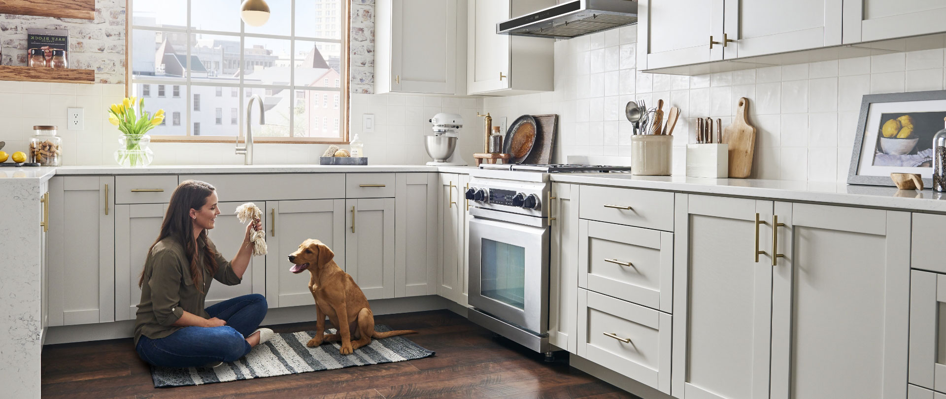 a woman sits on the floor in a kitchen playing with her dog